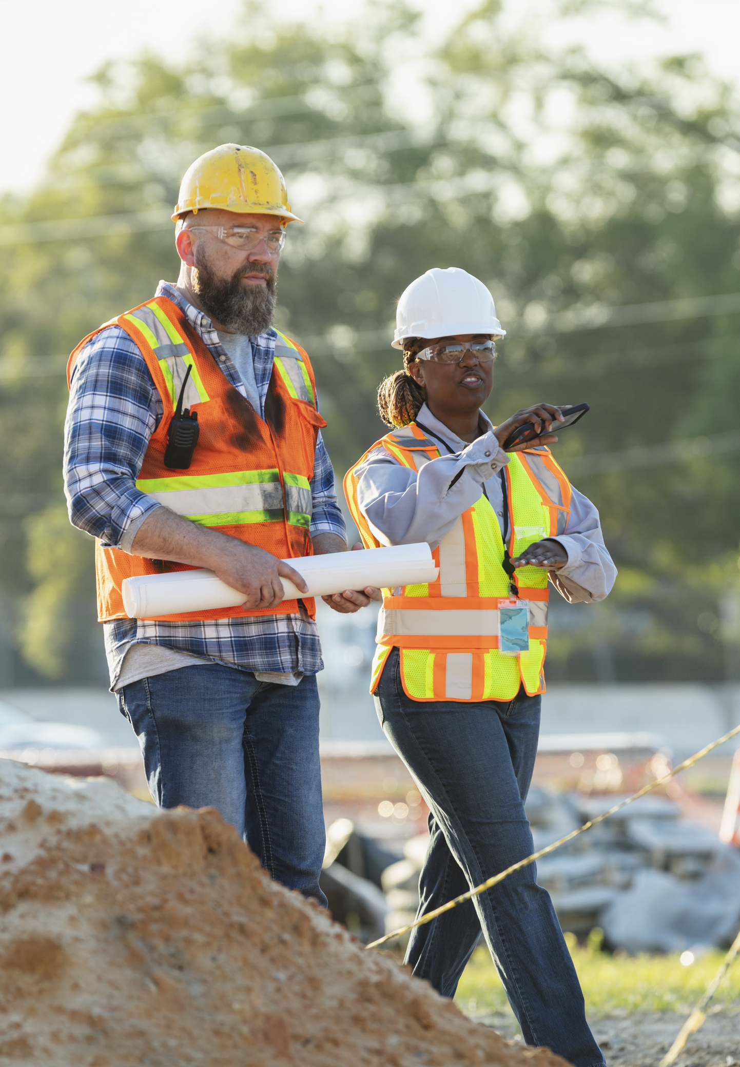 Two construction workers in safety vests and hard hats discussing project at a job site.
